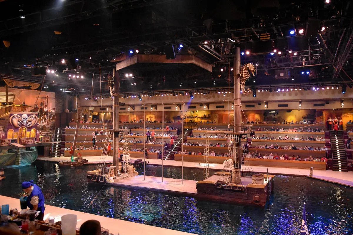 An energetic interior scene highlights a large pirate ship stage, a water-filled performance area, and excited audience seating, capturing the thrill of a live dinner show.