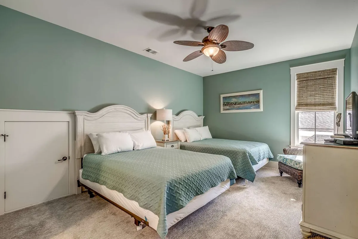 A spacious bedroom with two large beds, ceiling fan, window with shades, and a nightstand with a lamp. Subtle artwork adorns the wall above the beds, and the layout suggests comfort and flexibility for families or groups sharing the room.