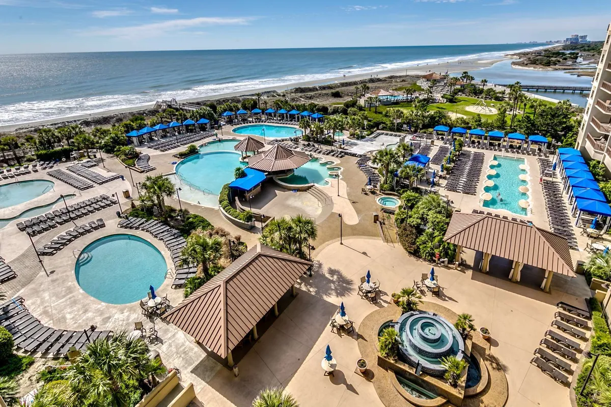 A sweeping exterior view showcases the resort’s expansive pool complex, featuring multiple sparkling pools surrounded by palm trees, lush landscaping, and inviting lounge areas. The beachfront location and scenic water features convey both relaxation and activity, hinting at the resort's many aquatic amenities.