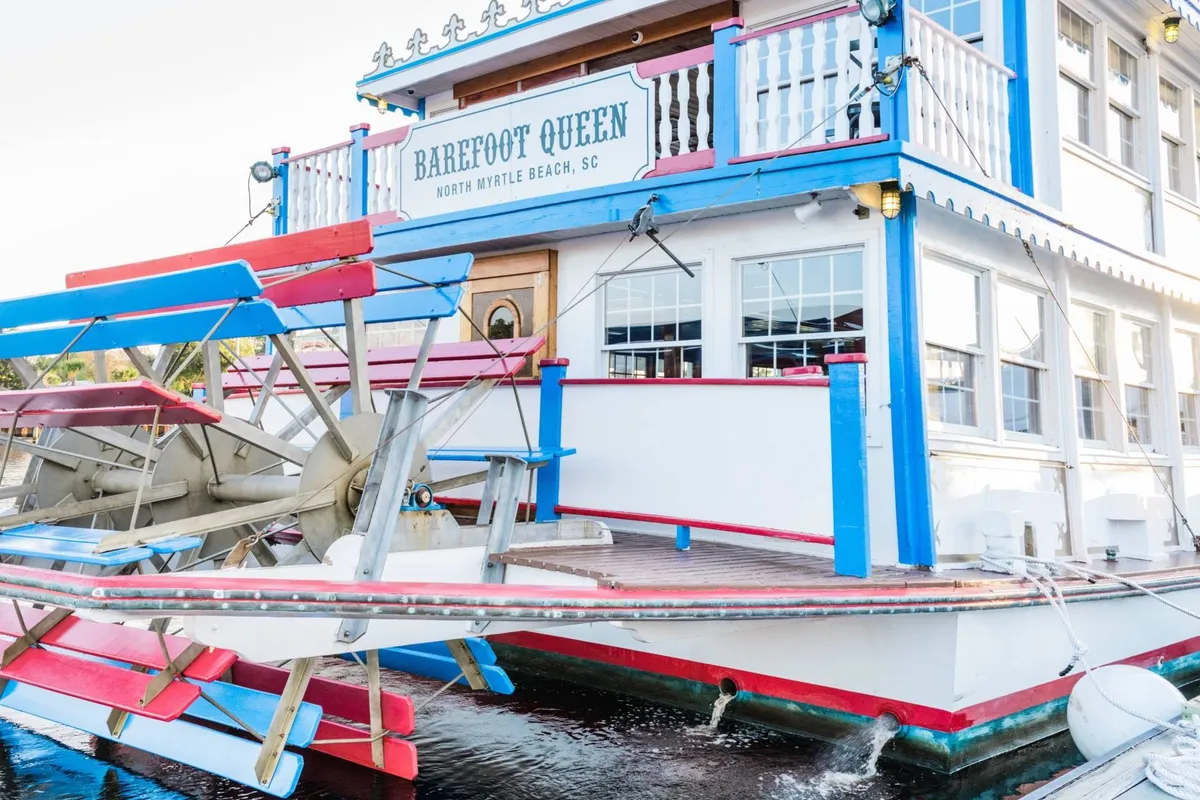 A lively exterior shot of the 'Barefoot Queen' paddlewheel boat, set against vibrant colors and the scenic waterway. This festive scene hints at unique dining and social experiences available across from the resort at Barefoot Landing.