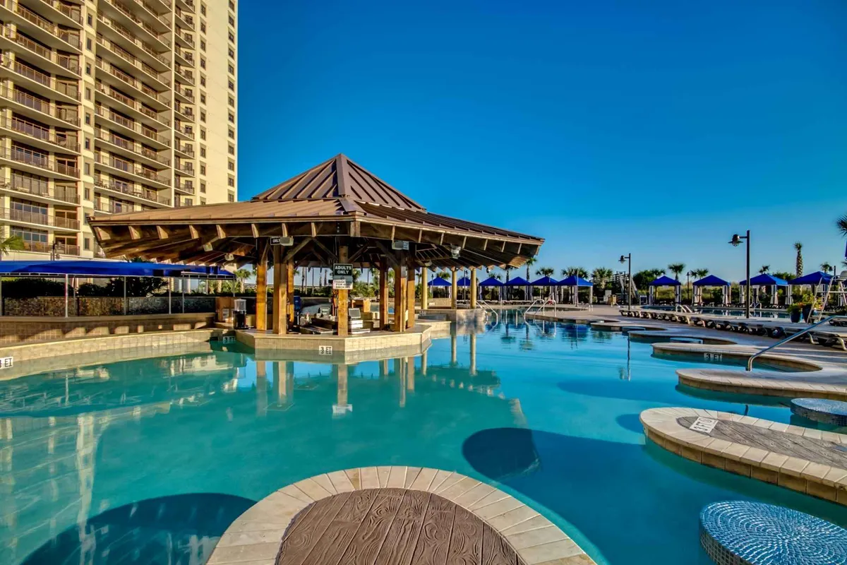 A vibrant pool scene with a shimmering pool, poolside cabana, numerous lounge chairs, and blue umbrellas. The area is bathed in sunlight, evoking a cheerful, social mood that invites guests to swim, sunbathe, or enjoy refreshing drinks by the cabana.