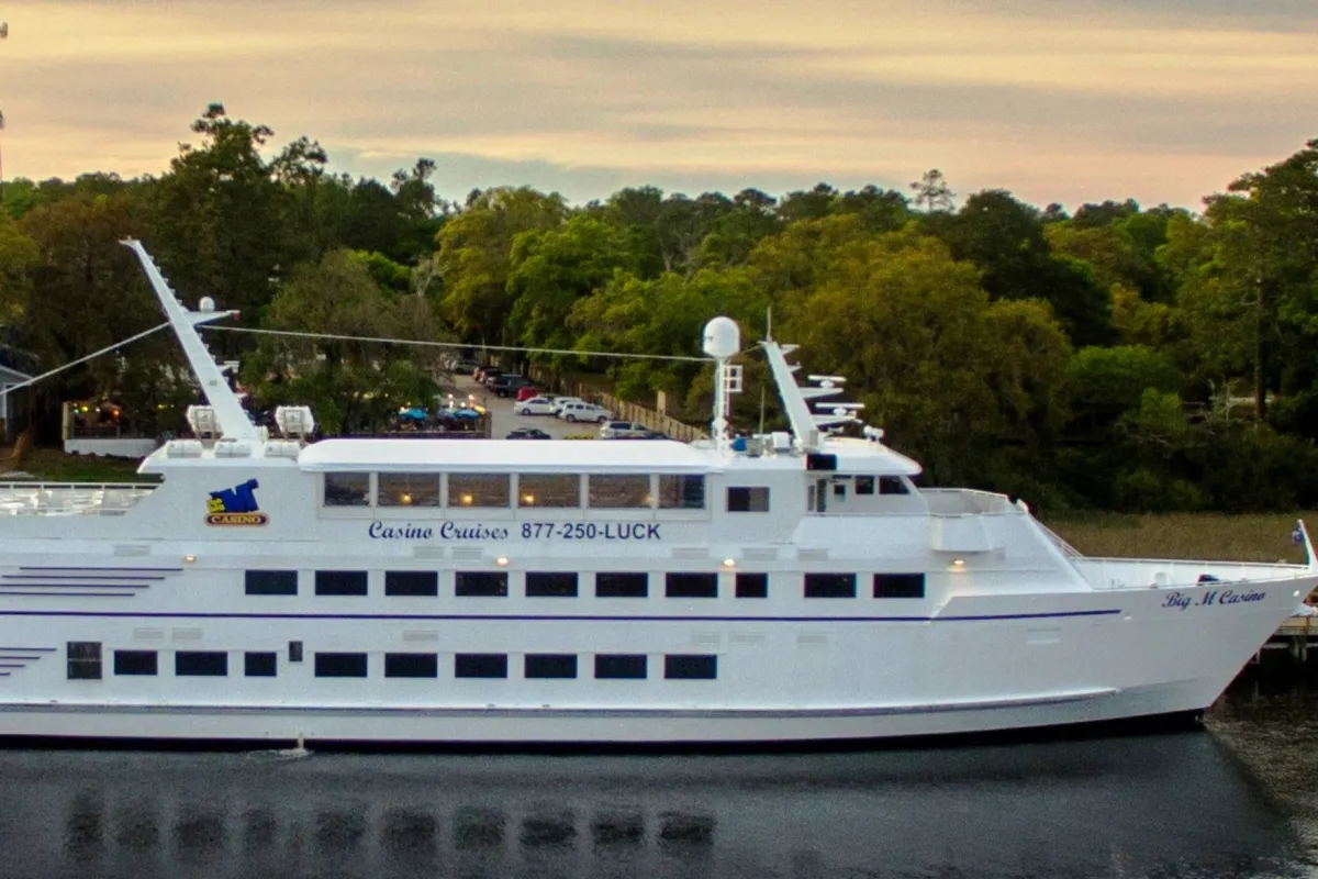 A large white casino cruise boat is docked amidst vibrant greenery, with the sky painted in warm sunset colors. The scene hints at social gatherings, sunset views, and exciting local experiences tied to the resort lifestyle.