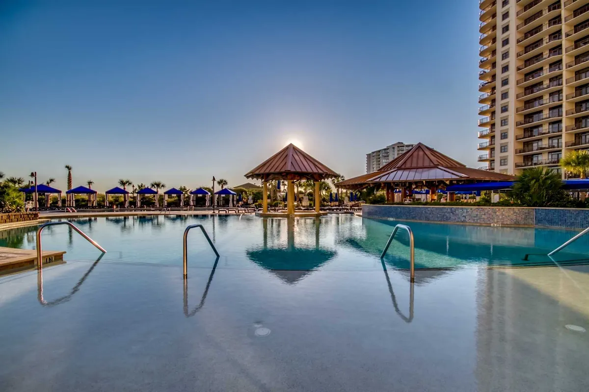 This image captures an inviting outdoor resort pool surrounded by palm trees and cabana structures. The water glistens as the sun sets, casting a golden glow across the scene, while lounge chairs suggest spaces for both relaxation and socializing.