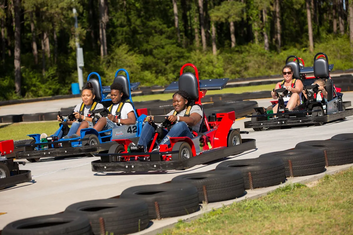 A lively go-kart race track with participants in action, surrounded by lush greenery. The image conveys the excitement and competitive spirit of family activities, with several people racing and others watching or waiting their turn.