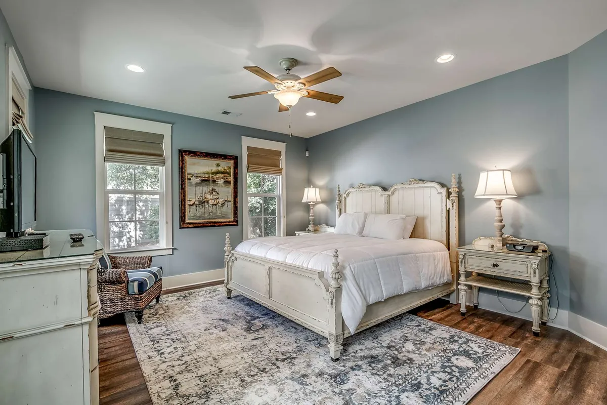 A serene bedroom featuring a neatly made bed with crisp linens, complemented by matching nightstands, a framed painting above the headboard, and a soft rug underfoot. Soft natural light filters gently into the space, inviting relaxation and comfort.